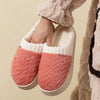 Pair of pink and cream plush fleece slippers being held, shown against a beige background.