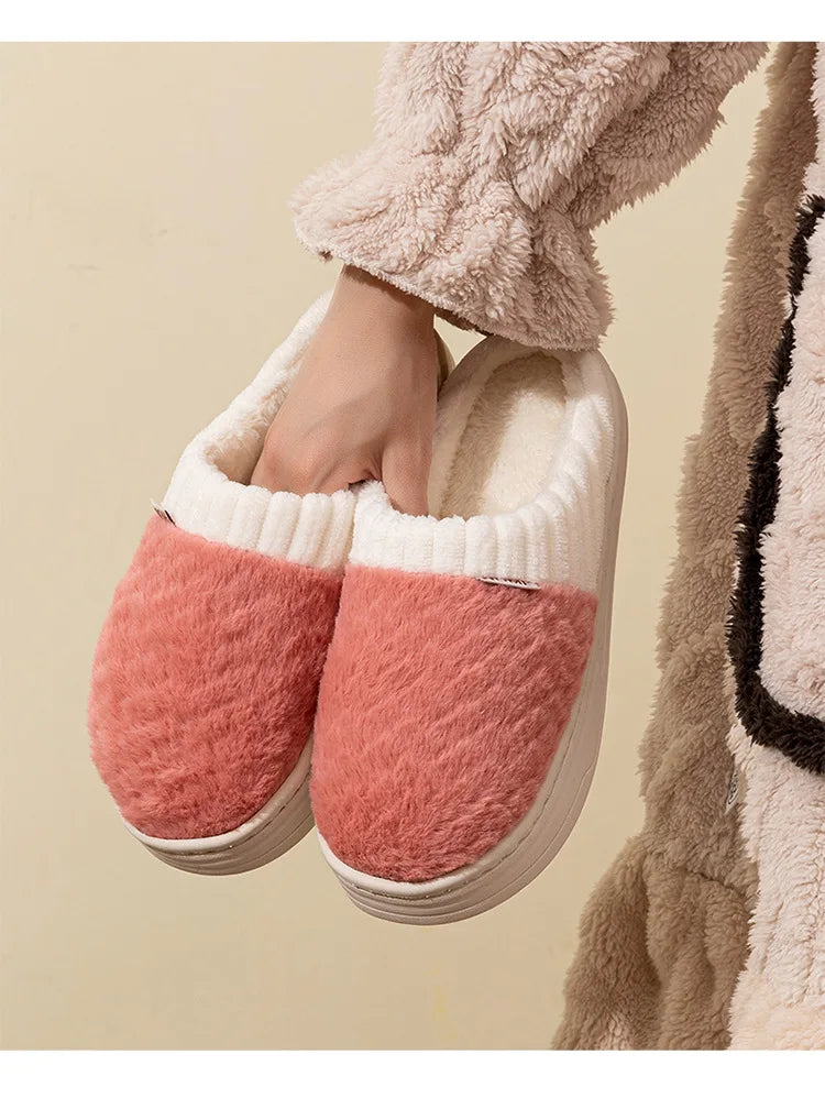 Pair of pink and cream plush fleece slippers being held, shown against a beige background.