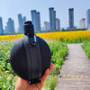 A hand holds a black collapsible silicone camping kettle outdoors with a city skyline in the background.