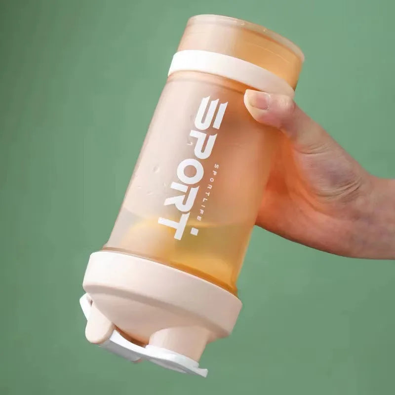 A hand holds a clear, orange-tinted 3-layer protein shaker bottle with a handle against a green background.