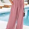 A model wearing dusty rose flowy high waist drawstring wide leg pants and a white cropped top standing by a pool.