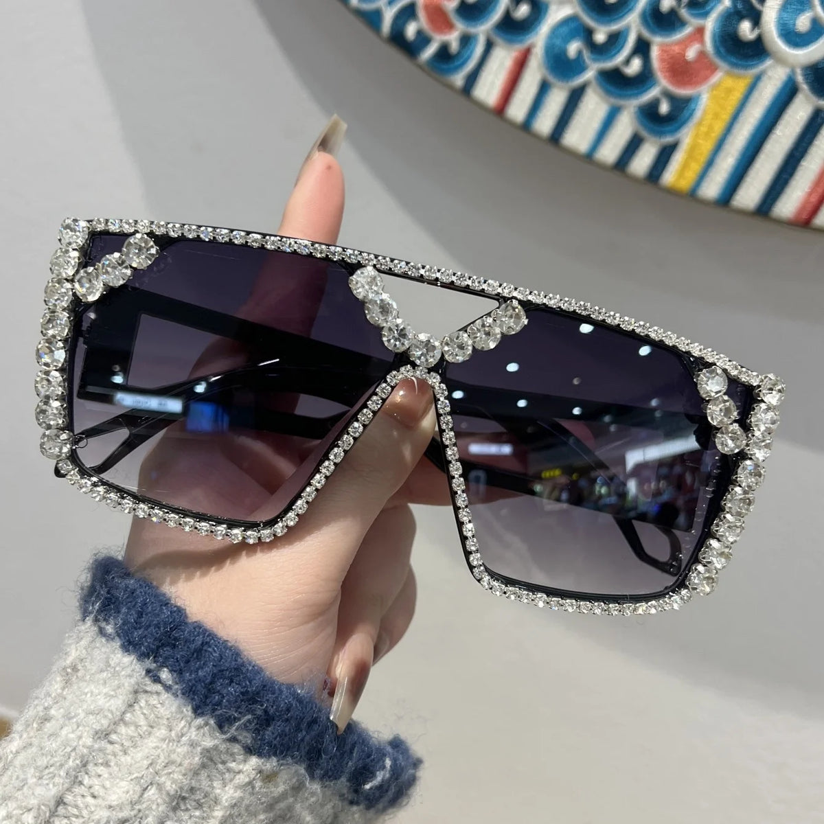 A hand holds a pair of black sunglasses decorated with clear rhinestones against a light background.