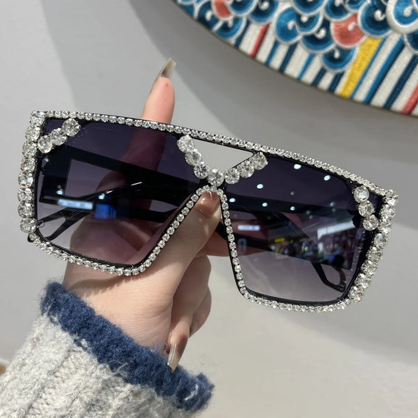 A hand holds a pair of black sunglasses decorated with clear rhinestones against a light background.