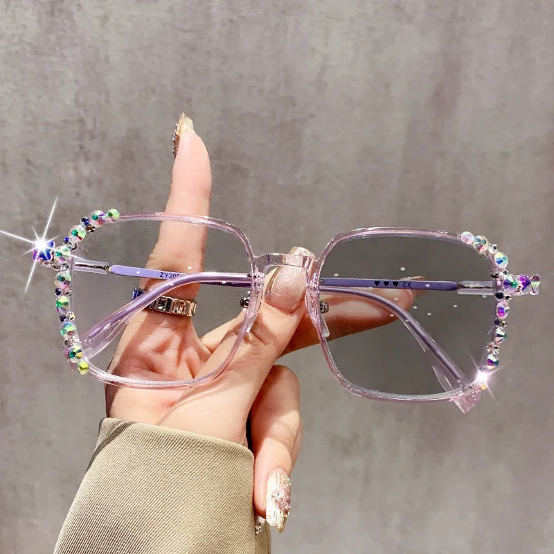 A hand holds a pair of pink photochromic diamond style eyeglasses with iridescent accents against a gray background.