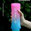 A hand holds a pink and blue inverted water bottle against a dark green, leafy background.