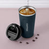 A dark blue insulated thermos bottle with a lid and a latte art coffee drink inside, resting on a pink surface.