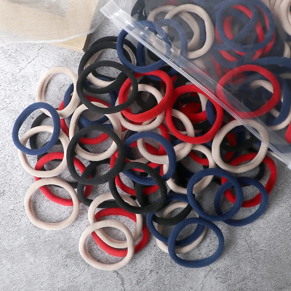 A pile of 50 mixed color elastic hair bands in black, red, blue, and beige on a clear plastic sheet.