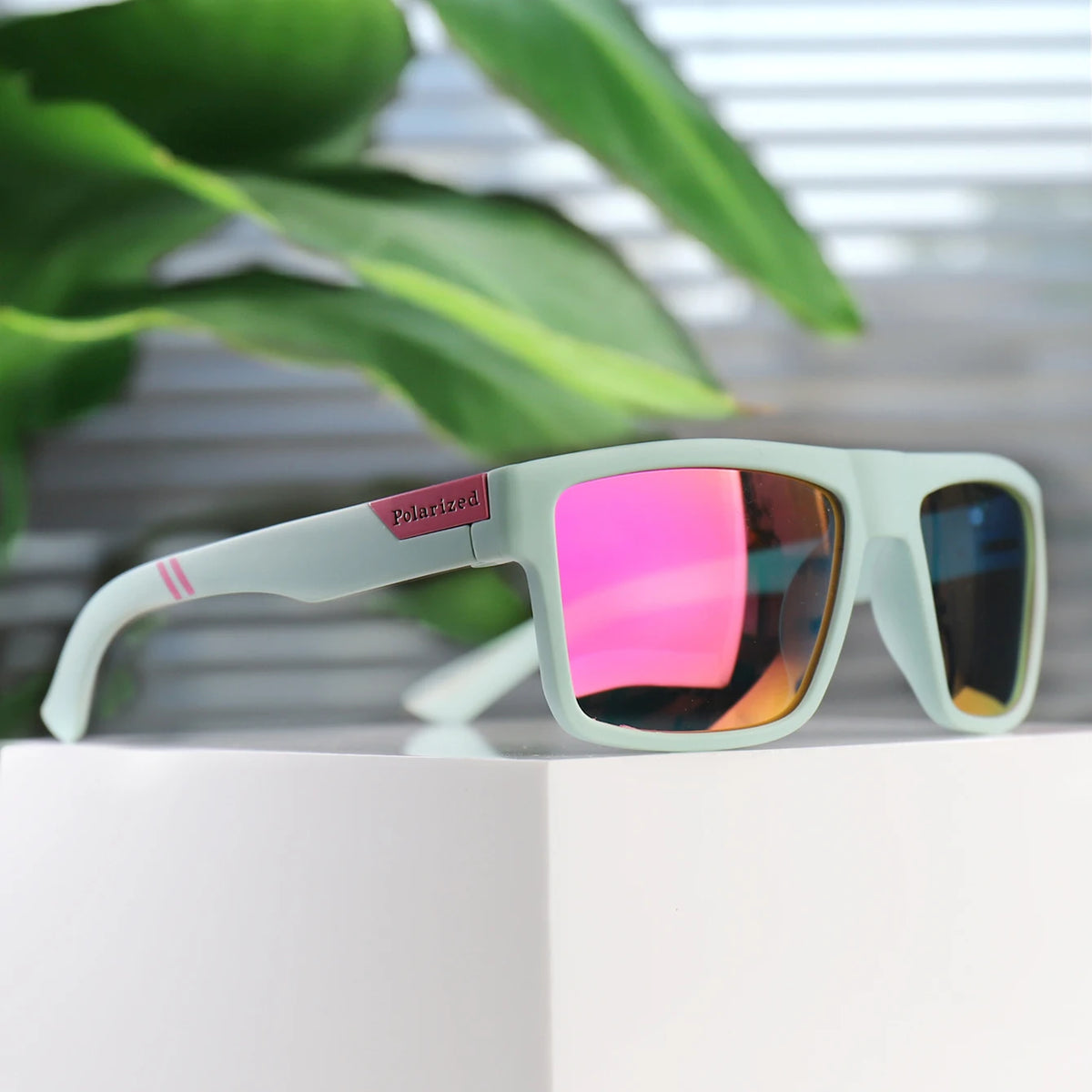 A pair of light green polarized square sunglasses resting on a white surface with blurred green foliage in the background.