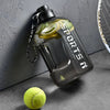 Klares Sportwasserflasche mit schwarzem Deckel und Limettenscheiben vor grauer Wand neben einem Tennisball.