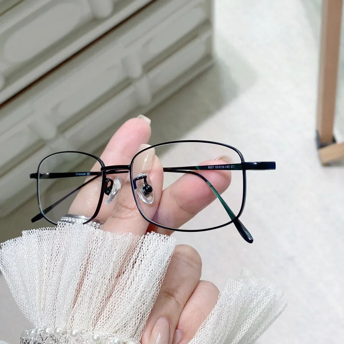 A hand holds a pair of black retro square eyeglasses with clear lenses against a white background.