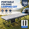 A portable folding camping cot with a padded mattress and steel frame is set up outdoors next to a tent.