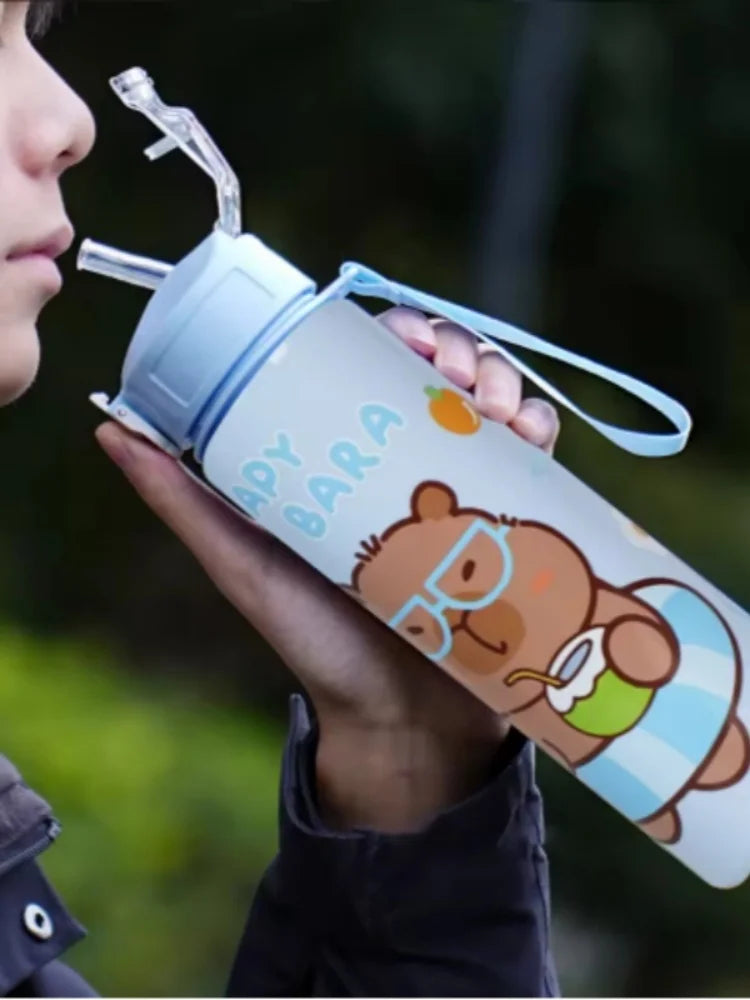 A person holds a cute capybara water bottle with a straw and handle against a blurred green background.