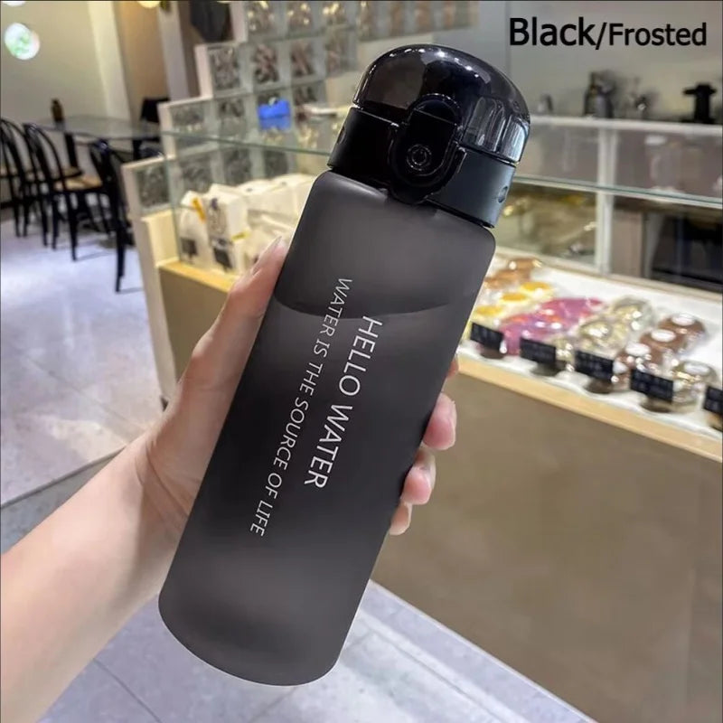 A hand holds a black frosted sports water bottle with a black cap, set against a blurred background of glass display shelves.