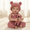 Baby in pink knitted bear hat and matching outfit sitting on a white blanket.