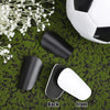 Black and white mini soccer shin guards displayed on green turf with a soccer ball in the background.
