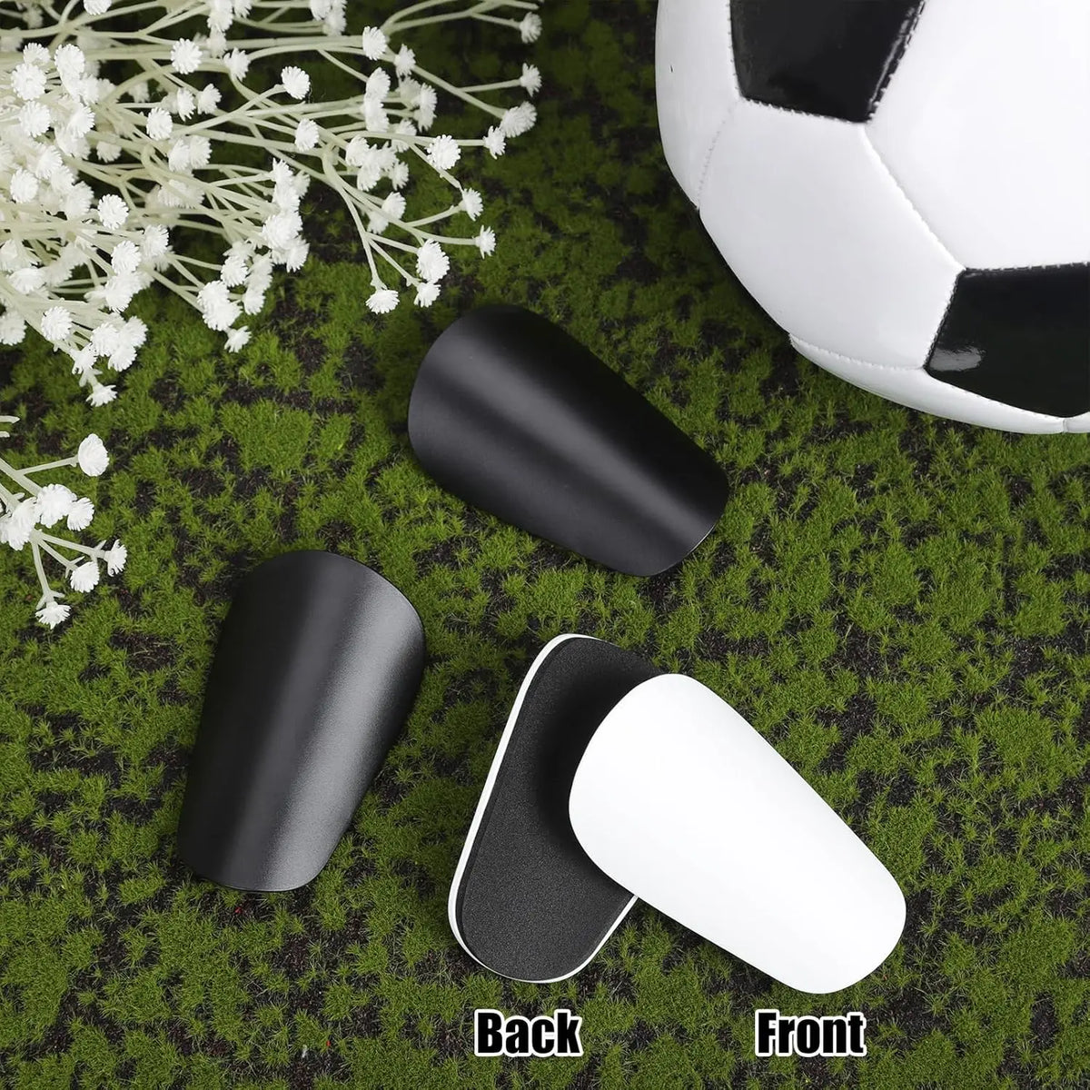 Black and white mini soccer shin guards displayed on green turf with a soccer ball in the background.
