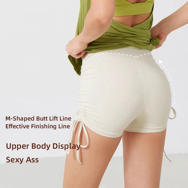 A woman wearing light beige performance yoga shorts with a green top, showcasing the M-shaped butt lift line.