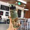 A hand holds a clear glass water bottle with a green cap and tea infuser on a wooden table.