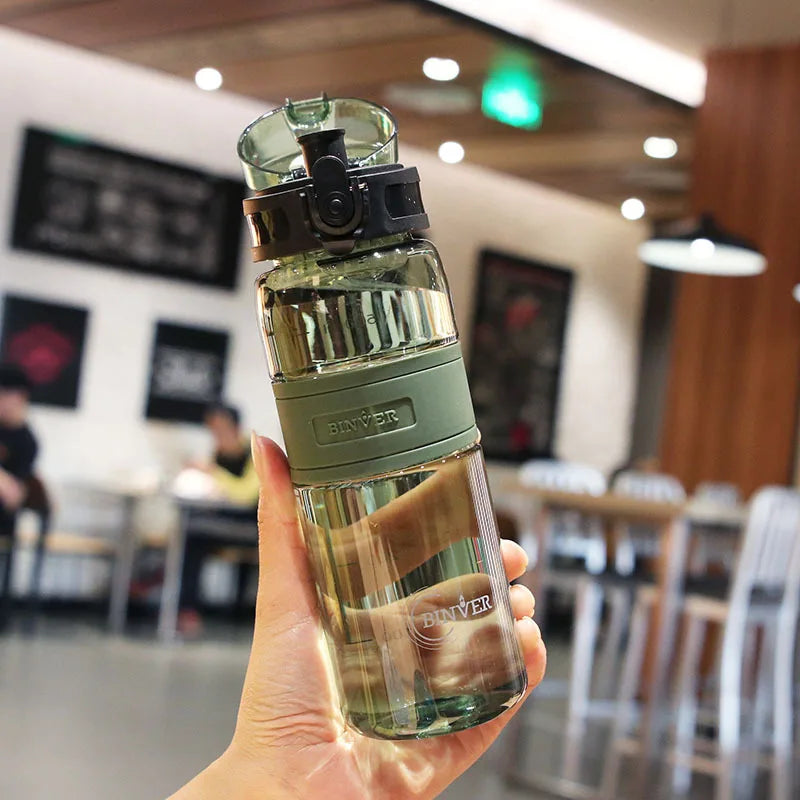 A hand holds a clear glass water bottle with a green cap and tea infuser on a wooden table.