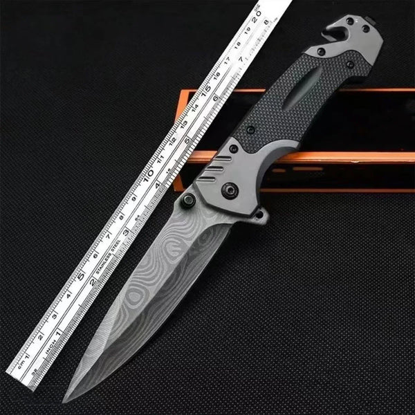 A multi-functional folding survival knife is shown next to a ruler for scale on a dark surface.