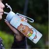 A hand holds a cute capybara water bottle with a straw and handle against a blurred green background.