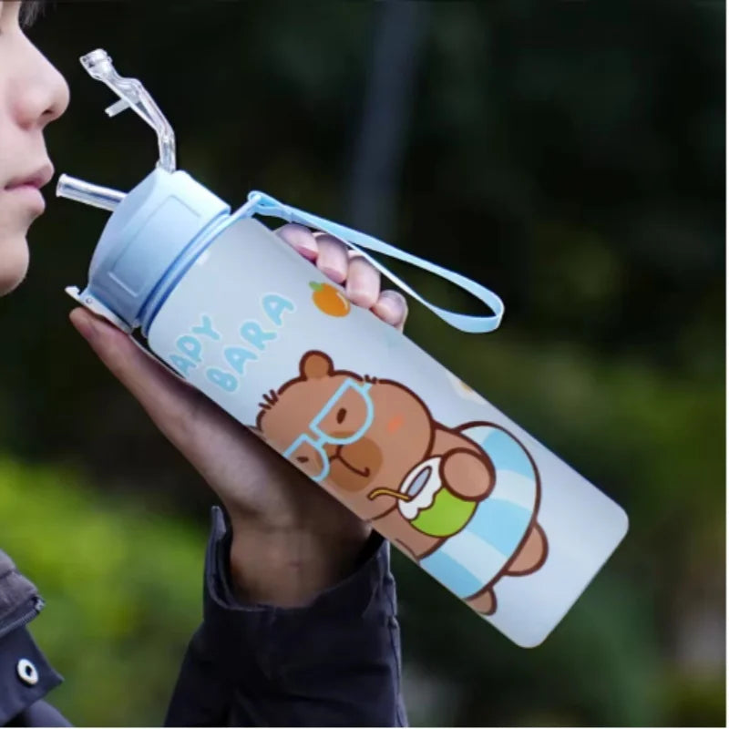 A hand holds a cute capybara water bottle with a straw and handle against a blurred green background.