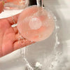 Une main tient un cache de téton en silicone transparent à centre perforé, lavé sous l'eau courante.