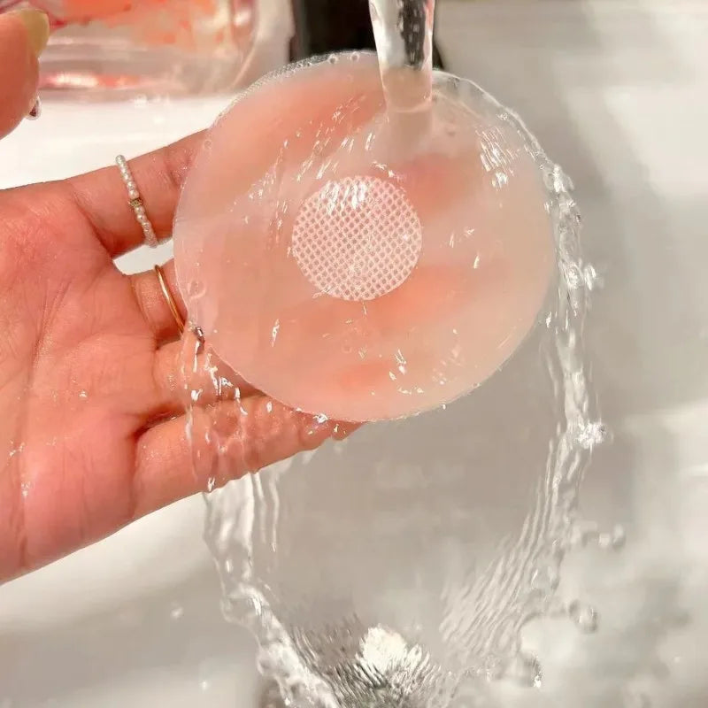 Une main tient un cache de téton en silicone transparent à centre perforé, lavé sous l'eau courante.