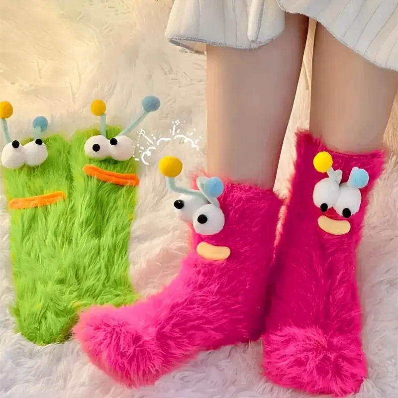 Pink fuzzy knit socks with cartoon expressions are shown on a white fluffy surface.