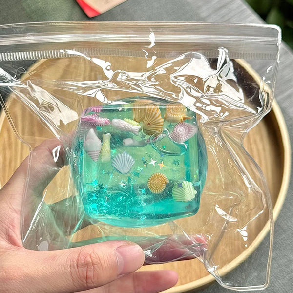 A hand holds a clear plastic bag containing a teal fidget cube with iridescent glitter and small shells.