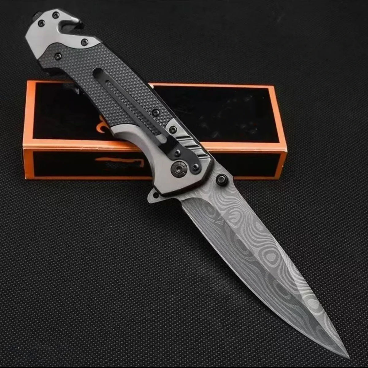A multi-functional folding survival knife with a silver blade and black handle is displayed on an orange box.