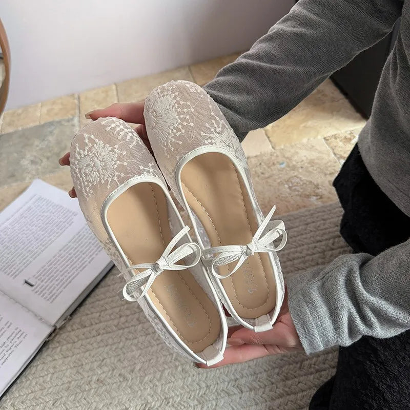 Una persona sostiene unas elegantes bailarinas de encaje con tela beige claro y cordones blancos.
