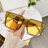 A hand holds a pair of oversized square yellow-tinted sunglasses with gold accents against a white background.