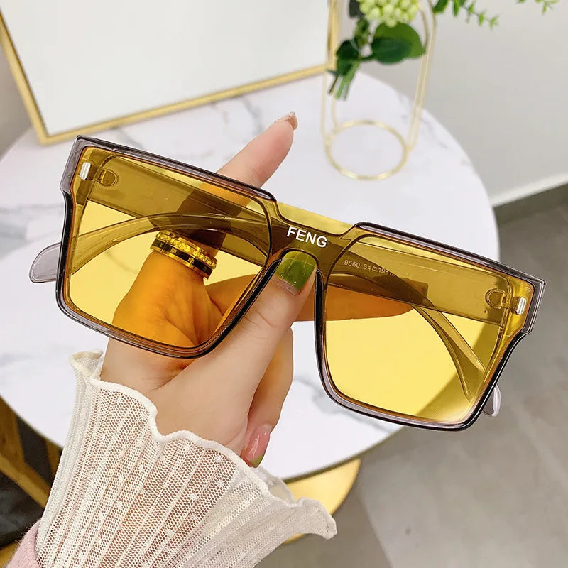 A hand holds a pair of oversized square yellow-tinted sunglasses with gold accents against a white background.