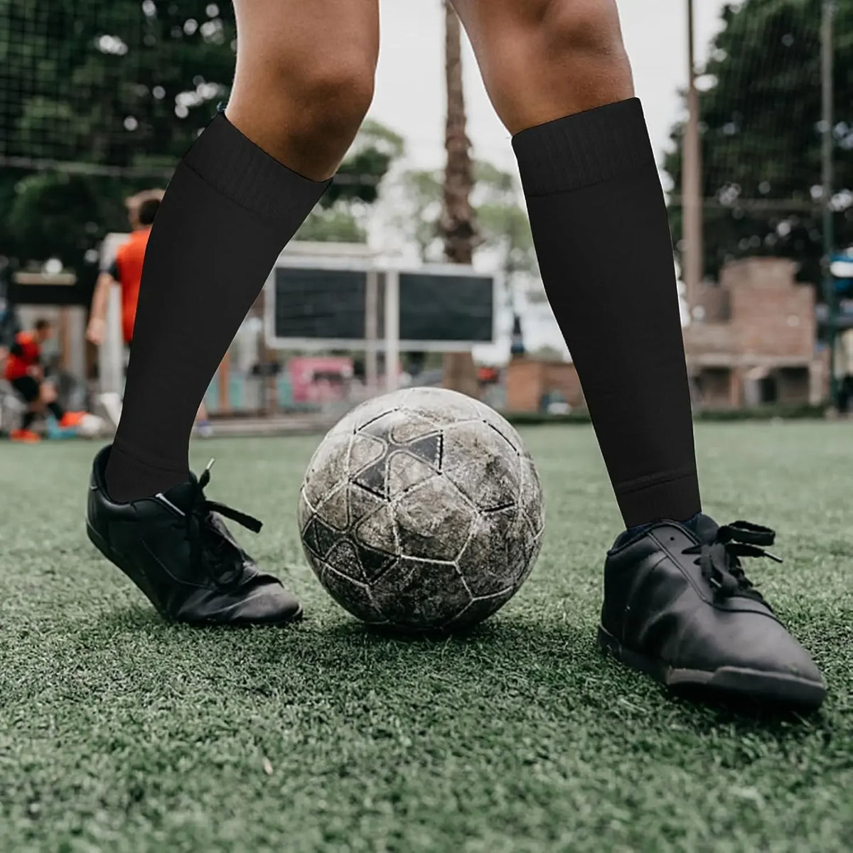 A person's legs wearing black compression sports socks and black cleats standing on a green field with a soccer ball.