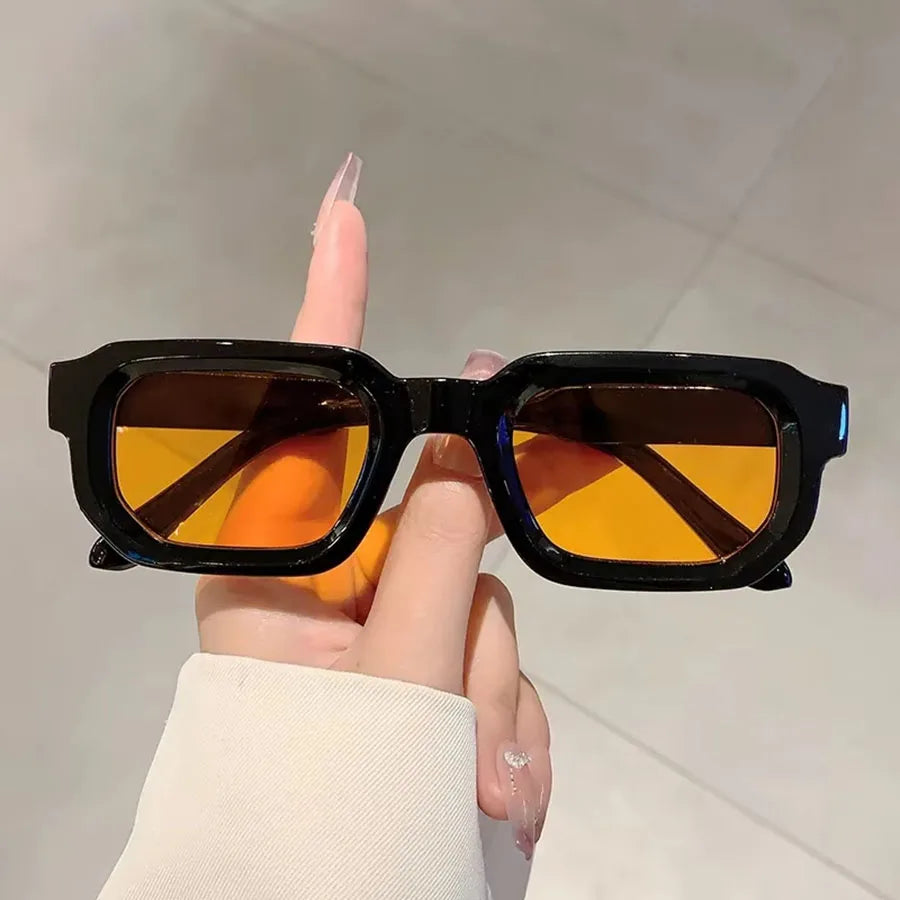 Une main tient des lunettes de soleil rectangulaires rétro noires avec des verres orange sur fond clair.
