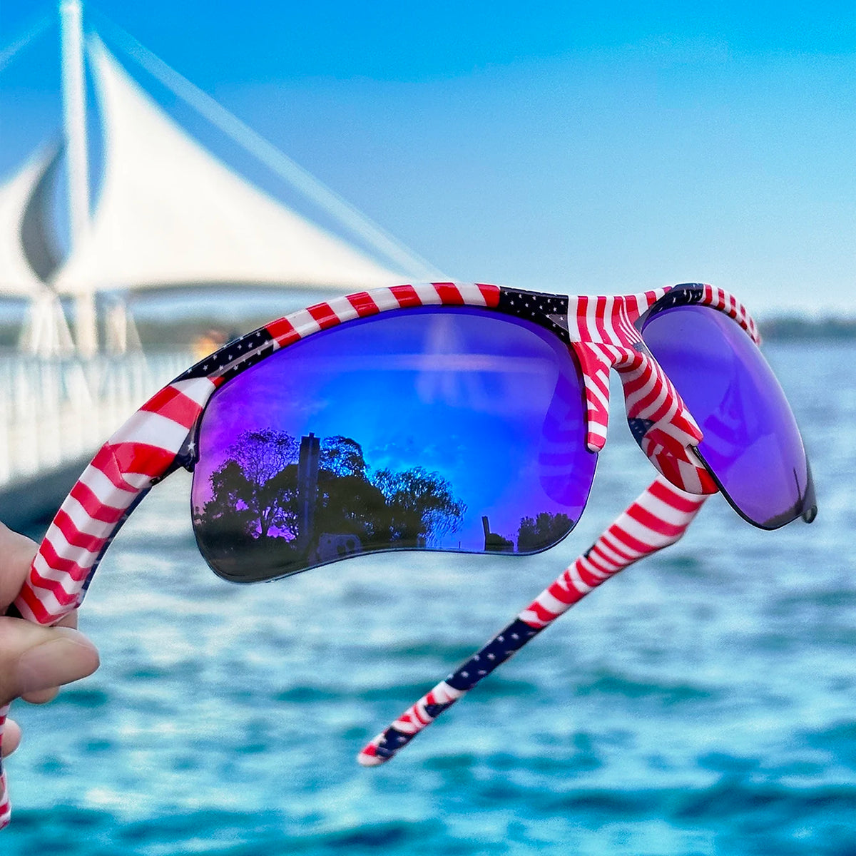 A person holds a pair of red, white, and blue striped polarized sports sunglasses against a bright blue sea background.