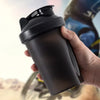 A hand holds a black portable protein shaker bottle against a blurred background.