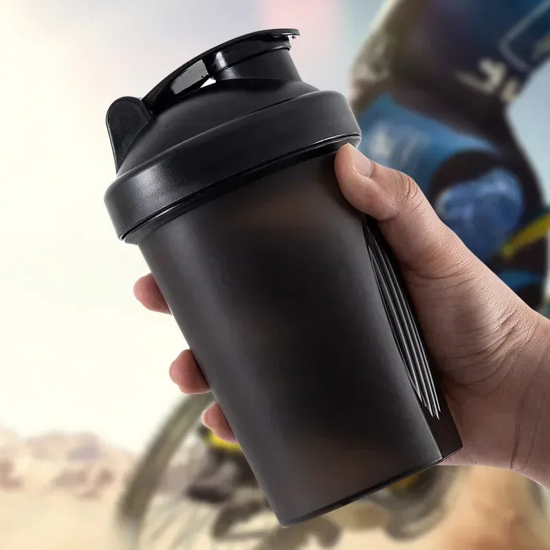 A hand holds a black portable protein shaker bottle against a blurred background.