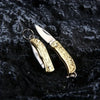 A brass folding knife keychain with an ornate handle and silver blade resting on a dark, textured surface.