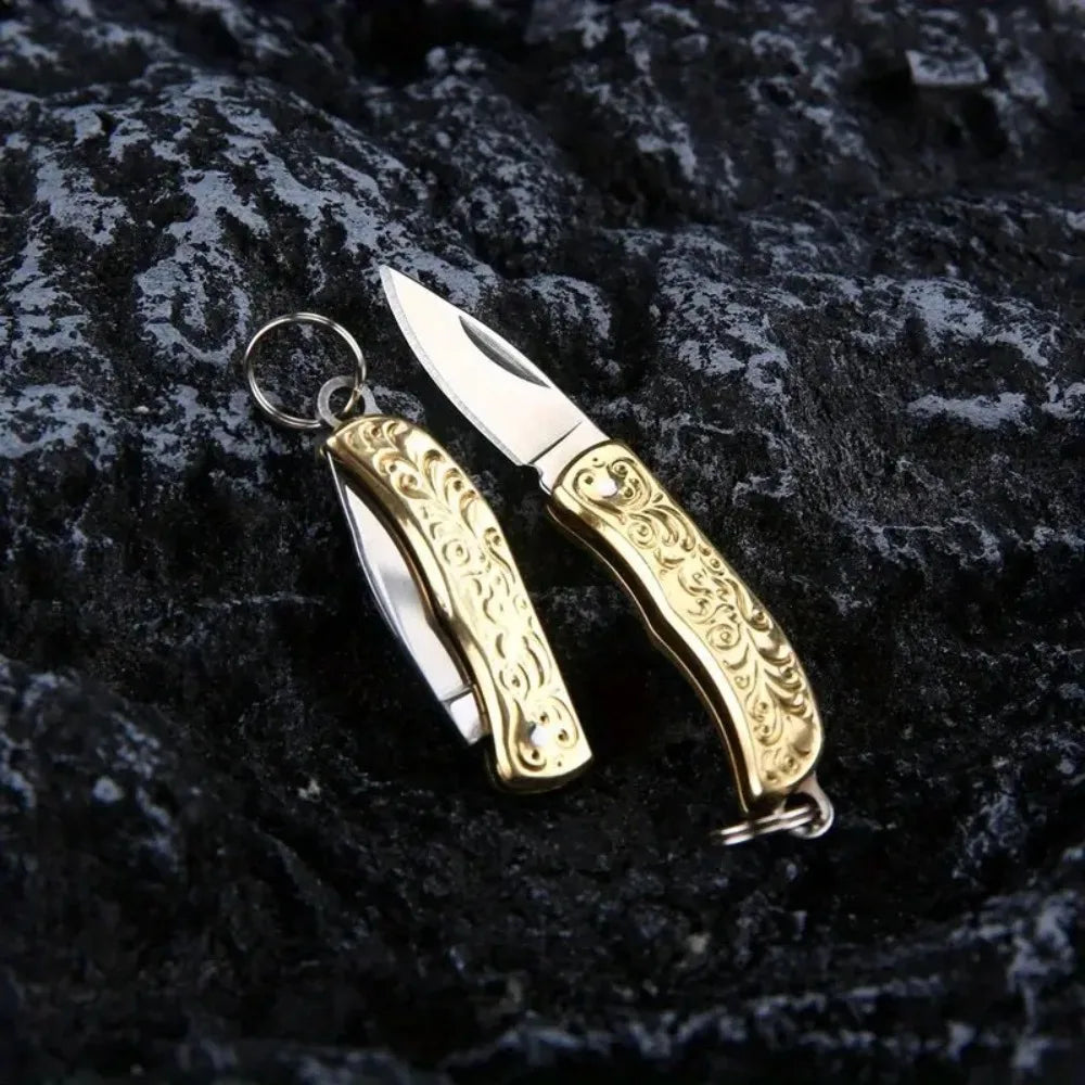 A brass folding knife keychain with an ornate handle and silver blade resting on a dark, textured surface.