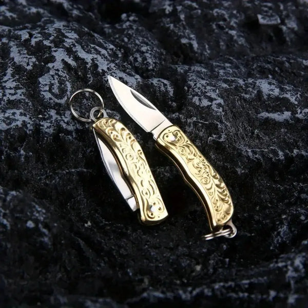 Couteau pliable en laiton avec poignée ornée et lame d'argent posé sur une surface sombre et texturée.