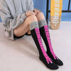Une personne porte des chaussettes noires montantes avec des rayures roses et des poignets gris, assise sur une surface blanc