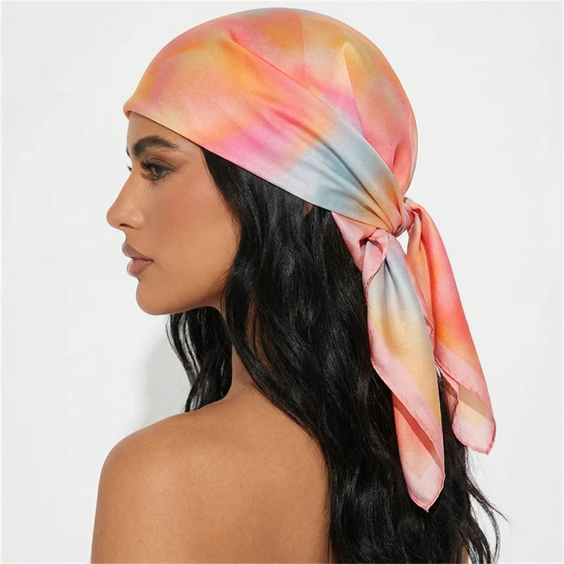 A woman wearing a silk-touch floral hair scarf tied around her head, featuring soft pink and orange watercolor tones.