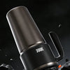 A close-up shot of a matte black insulated stainless steel tumbler with a handle and straw resting on rough, dark terrain.