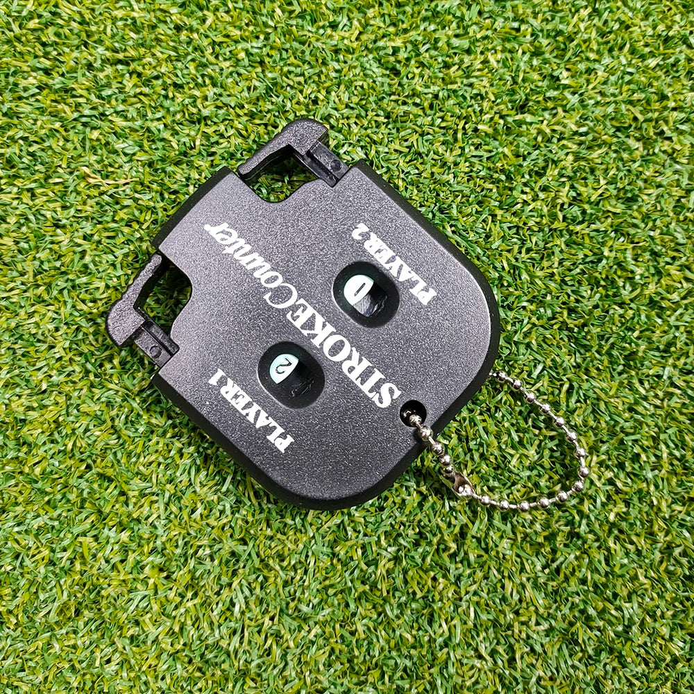 A black portable golf score counter device with a key ring attached, resting on green grass.