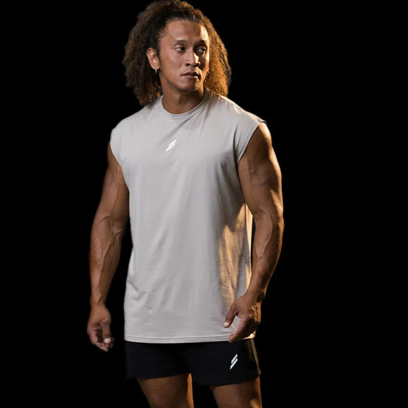 A man wearing a light gray sleeveless fitness tank and black shorts posing against a black background.