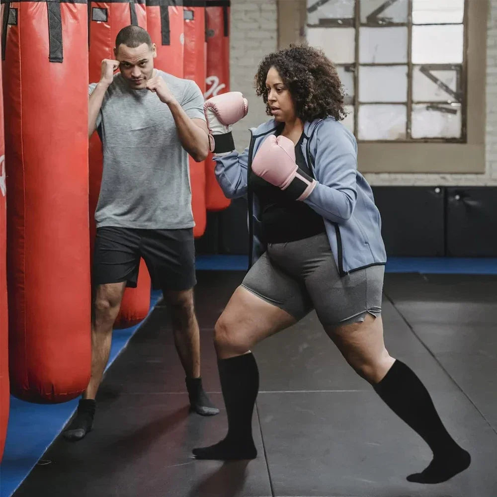 A woman wearing a gray shirt and black athletic wear is training with a man in a gym setting.