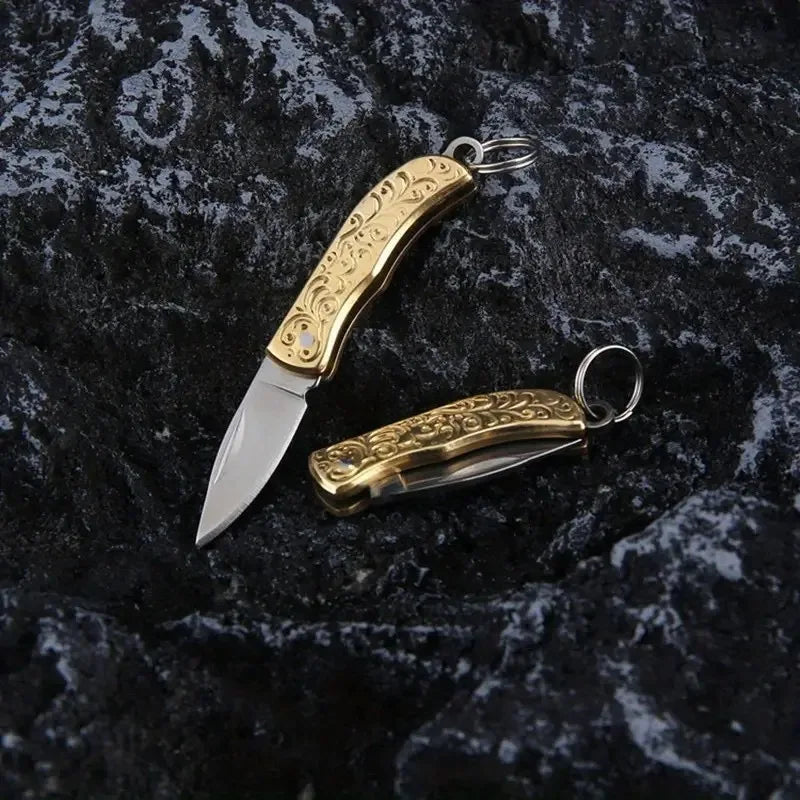 A brass folding knife keychain with an ornate handle and blade is displayed on a dark, textured surface.