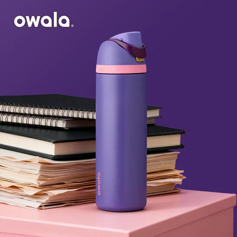 A purple Owala insulated stainless steel tumbler standing upright on a pink surface next to stacks of papers.